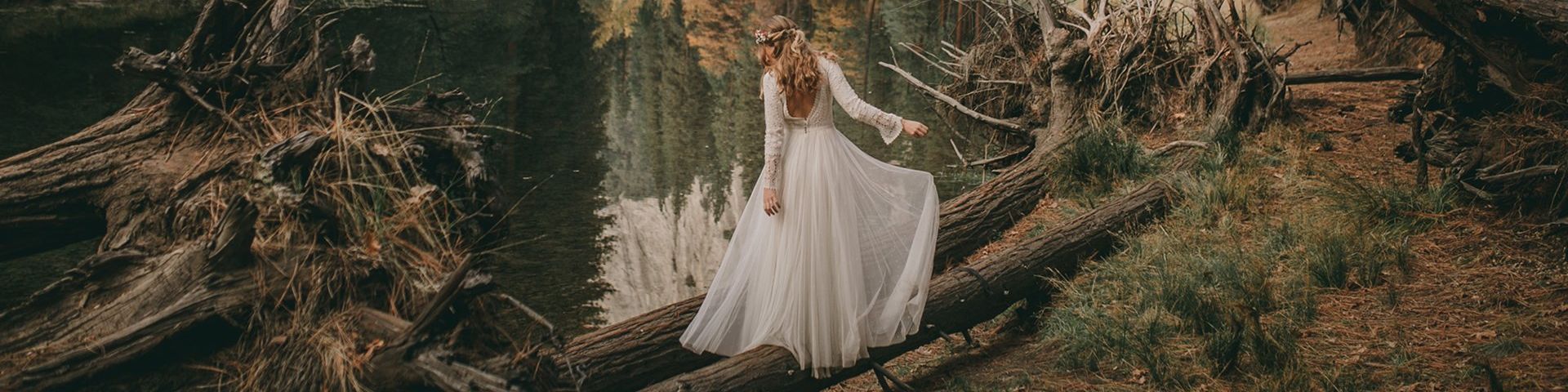 A women with long hair in her wedding dress is balancing on fallen trees on the edge of a give in a forest.